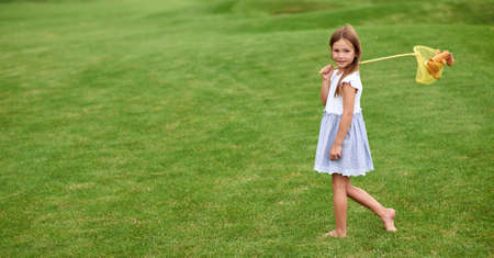 Full length shot of adorable little girl looking at camera, standing with a butterfly net, catching butterflies in the green park on a summer dayの写真素材