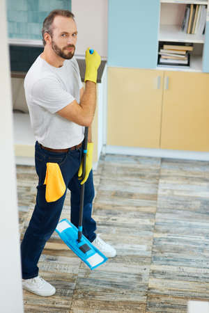 Making your house clean. Full length shot of a man, professional male cleaner wearing gloves looking at camera, holding mop while cleaning floor in the kitchenの写真素材