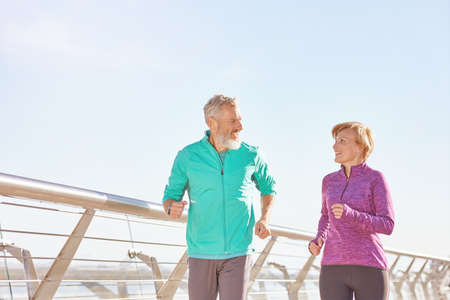 Energize your life. Active mature family couple in sportswear smiling at each other while running together on a sunny morning. Joyful senior couple working out outdoorsの写真素材