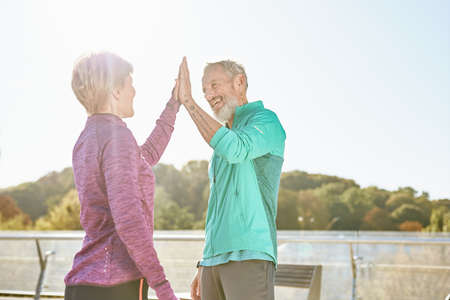 Partnership. Happy family couple, mature man and woman in sportswear giving high five after having workout in the city park on a sunny morningの写真素材