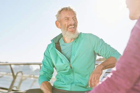 Cheerful sporty senior couple resting, sitting on the bench after workout outdoors. Mature man smiling to woman while discussing somethingの写真素材