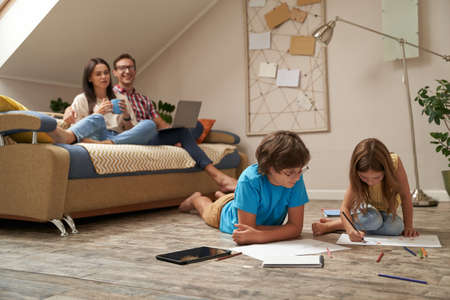 Happy mother and father sitting on sofa and watching movie on laptop while their kids, little boy and girl lying on the floor and drawing with colorful pencilsの写真素材