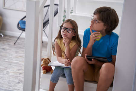 Smart kids. Thoughtful little brother and sister sitting on stairs at home and doing homework together, learning onlineの写真素材