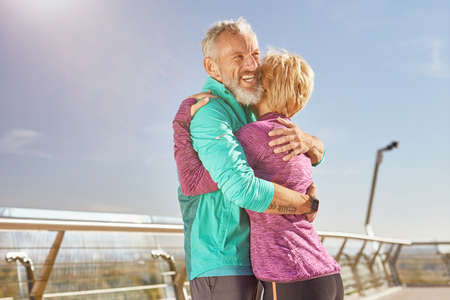 Support. Happy mature family couple in sportswear smiling while hugging after having workout in the city park on a sunny morning. Joyful senior couple working out together outdoorsの写真素材