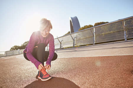 Get Ready. Full length shot of active mature woman wearing sportswear tying shoelaces on sporty sneakers while getting ready for running outdoors on a sunny dayの写真素材