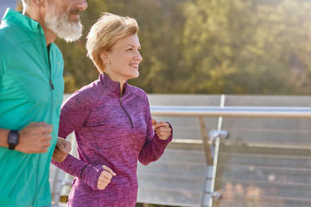 In motion. Cheerful active mature woman in sportswear smiling while running on a sunny day. Joyful senior couple doing sport together outdoorsの写真素材