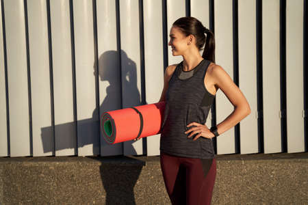 Smiling pretty lady holding exercise mat outdoorsの写真素材