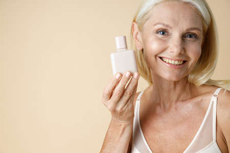 Close up portrait of gorgeous mature blonde woman in underwear smiling at camera, holding white bottle of cream or lotion for skin care, posing isolated over beige backgroundの写真素材