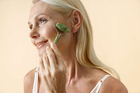 Close up portrait of mature blonde woman with glowing skin smiling aside while using jade facial roller for skin care and beauty treatment, posing isolated over beige backgroundの写真素材