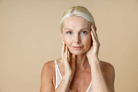 Close up portrait of charming mature woman with blonde hair in white underwear looking at camera, touching her face isolated over beige backgroundの写真素材
