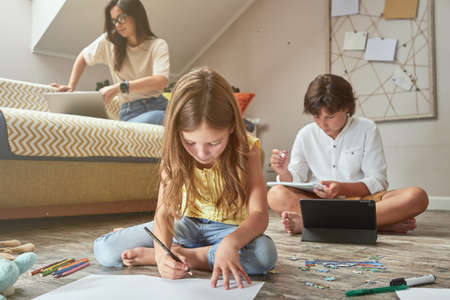 Little girl sitting on the floor and drawing while her brother doing homework online on digital tablet, young beautiful mother sitting on sofa with laptop on backgroundの写真素材