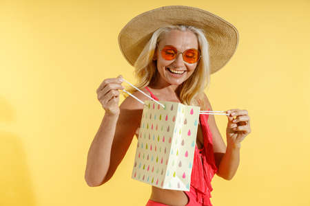 Excited fashionable woman in red swimsuit and straw hat looking surprised while opening and looking inside shopping bag, standing isolated on yellow backgroundの写真素材