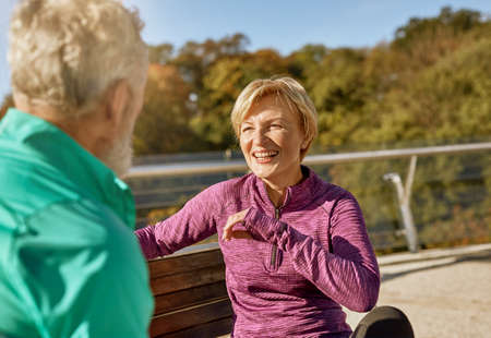 Happy sporty senior couple resting, sitting on the bench after workout outdoors. Mature woman smiling to man while discussing somethingの写真素材
