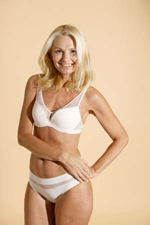 Cheerful caucasian mature blonde woman in lingerie smiling at camera, standing isolated over beige background, posing half nakedの写真素材