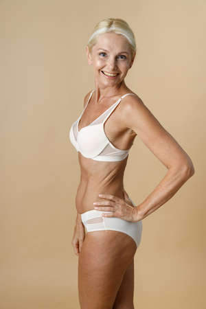 Pretty mature woman in underwear with fit body turning head to camera and smiling, holding hand on waist standing isolated over beige backgroundの写真素材