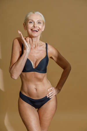 Studio shot of excited mature woman in black underwear smiling at camera while standing and posing half naked isolated over beige backgroundの写真素材