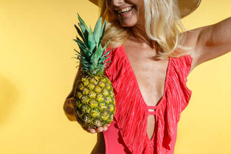 Excited blonde woman in red swimsuit and hat smiling, holding fresh ripe pineapple fruit while posing isolated on yellow background in studio. Focus on pineappleの写真素材