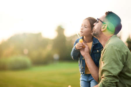 Adorable little girl having fun with soap bubbles on a sunny day, daughter and dad spending time together outdoorsの写真素材