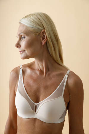 Portrait of beautiful mature blonde woman in white bra posing with eyes closed isolated over beige backgroundの写真素材