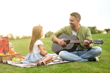 Cute little girl in casual clothes sitting on a green grass in park and looking at her loving dad playing guitarの写真素材