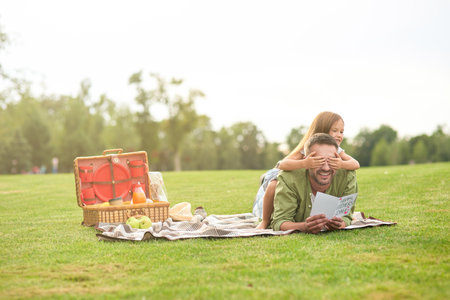 Full length shot of cute little girl covering fathers eyes while greeting and giving him handmade postcard, daddy and daughter having a picnic in the parkの写真素材