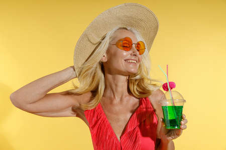 Relaxed blonde woman in sunglasses, red swimsuit and straw hat smiling with eyes closed, adjusting her hat and holding cocktail drink while posing isolated on yellow backgroundの写真素材
