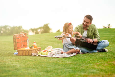 Full length shot of happy little girl sitting with her loving father on a green grass in park and learning how to play guitarの写真素材