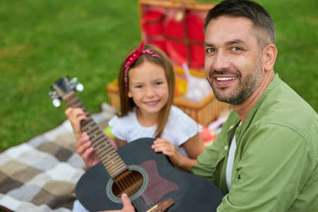 Portrait of happy father smiling at camera while sitting with his lovely daughter on green grass in park and holding guitarの写真素材