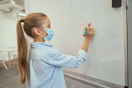Intelligent little schoolgirl wearing protective mask during coronavirus pandemic writing numbers on...