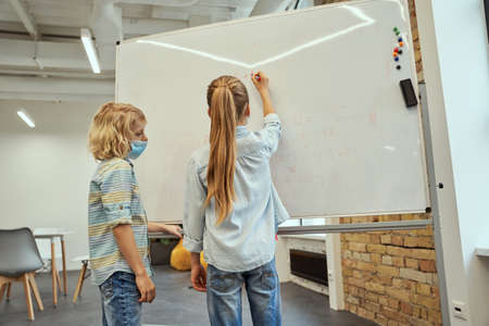 Little school kids, boy and girl wearing protective mask during coronavirus pandemic writing on...