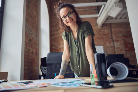 Creative woman, interior designer or architect in casual wear with messy hairdo smiling at camera while working on a blueprint for new project, standing in her officeの写真素材