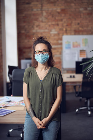 Creative young woman in casual wear wearing glasses and protective mask looking at camera, leaning on the table while standing in modern office on a daytimeの写真素材