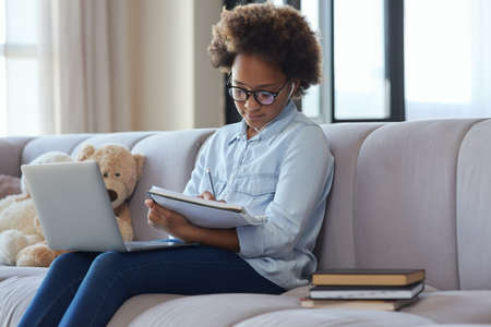 School at home. Teen schoolgirl wearing earphones making notes in her notebook while studying using laptop, sitting on the couch at homeの写真素材