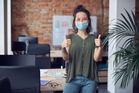Cheerful creative woman wearing protective mask smiling at camera, showing thums up while standing in modern green office on a daytimeの写真素材