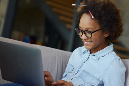 Successful student. Joyful teen schoolgirl in glasses with pencils in her hair smiling while using laptop for studying, sitting on the couch at homeの写真素材