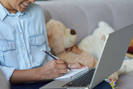 Cropped shot of cheerful teen schoolgirl using laptop, making notes while sitting on the sofa at homeの写真素材
