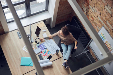 High angle view of female interior designer taking headphones, reading a blueprint while working on new project, sitting in modern officeの写真素材