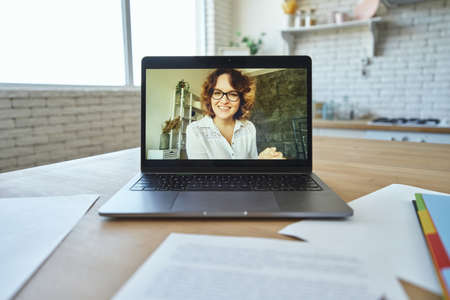 Attractive female teacher smiling at camera during video call, having online lesson with students studying from home. Focus on laptop screenの写真素材