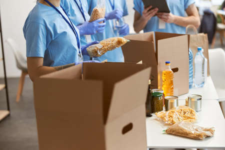 Cropped shot of volunteers in protective gloves sorting, packing food in cardboard boxes, working on...