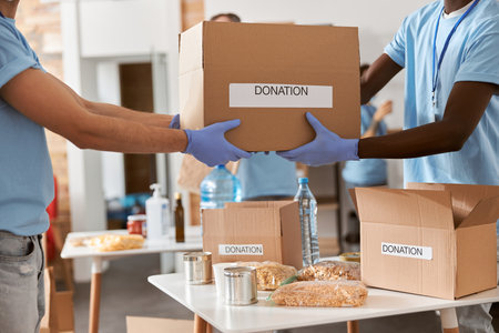 Cropped shot of diverse male volunteers in blue uniform, protective mask and gloves carrying cardboard box with donated food stuffs while standing indoorsの写真素材