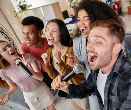 Face the song. Enthusiastic multicultured friends singing with microphone while playing karaoke at home, standing in the modern apartmentの写真素材