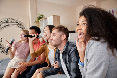 In love with karaoke. Group of young friends having fun, singing with microphone while playing karaoke at home, sitting on the couch in the modern apartmentの写真素材