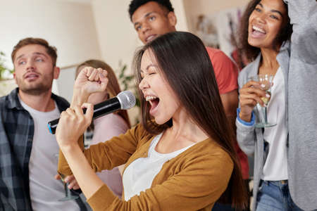 Perform like a superstar. Pretty young asian girl holding microphone and singing while playing karaoke with best friends at home, group of people drinking and having fun together. Selective focusの写真素材