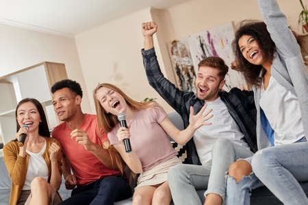 Feel the music. Group of multicultured friends looking excited while singing with microphone, playing karaoke at home, sitting on the couch in the modern apartmentの写真素材
