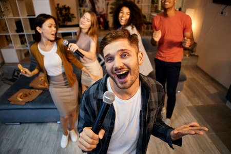 Show your singing skills. Happy young guy singing with microphone while playing karaoke with friends...
