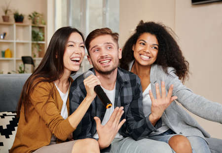 Karaoke singers. Three smiling friends looking happy while playing karaoke at home, singing with microphone, sitting on the couch in the modern apartmentの写真素材