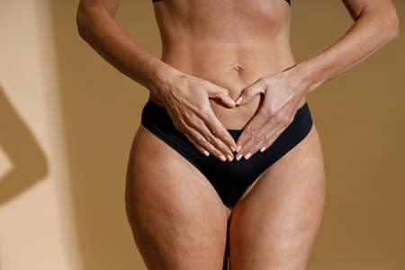 Close up shot of womans hands forming a heart symbol on belly. Perfect fit body of mature woman in black underwear holding hands on her stomach while standing isolated over beige backgroundの写真素材
