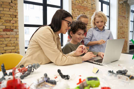 Attractive young female teacher in glasses showing scientific robotics video to curious kids using...