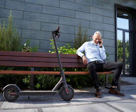 Full length shot of confident, cheerful middle aged businessman smiling while talking on the phone, sitting on the bench outdoors near modern buildingの写真素材