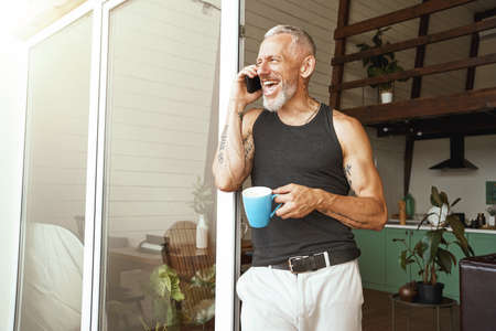 Enjoying good talk. Happy middle aged caucasian man talking by mobile phone and laughing while drinking coffee near balcony of his modern flatの写真素材